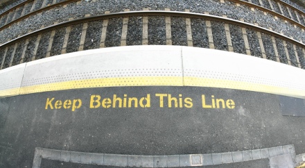 Peron, brzeg peronu i tory kolejowe. Zółta linia biegnąca wzdłuż torów po peronie. Żółty napis mówiący po angielsku 'Trzymaj się za linią', tylko że napis jest umieszczony przed linią.