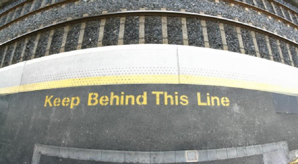 Peron, brzeg peronu i tory kolejowe. Zółta linia biegnąca wzdłuż torów po peronie. Żółty napis mówiący po angielsku 'Trzymaj się za linią', tylko że napis jest umieszczony przed linią.